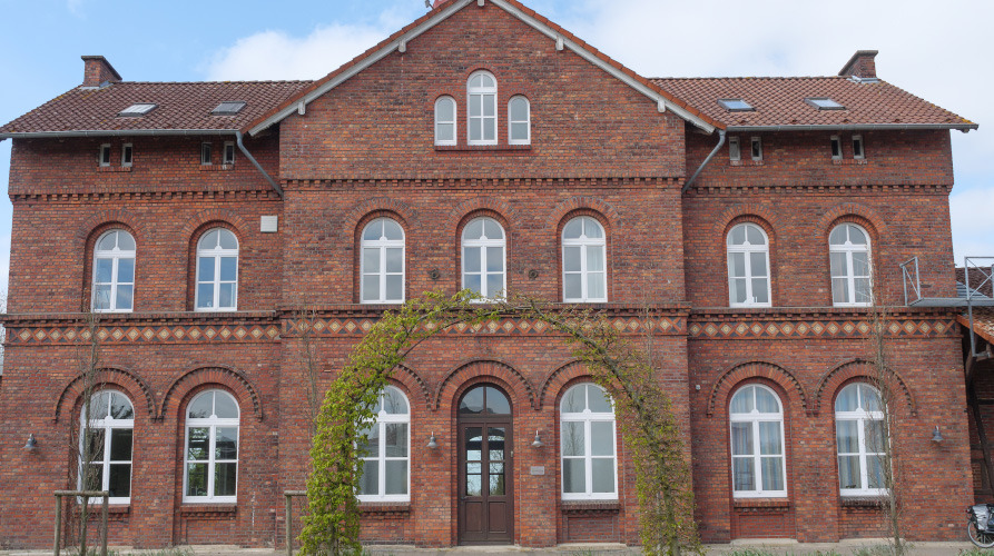 Ein großes, Gebäude aus rotem Backstein mit weißem Rundbogenfenster, einer zentralen Türe und einem grünen Bogen mit Kletterpflanzen an der Vorderseite. 