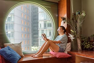 Frau sitzt an einem Rundfenster und liest ein Buch inmitten von Kissen und Pflanzen. Und das natürliche Licht genießt, das durch das runde Fenster hereinströmt.