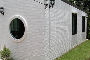 Ein kleines, weißes Backsteingebäude mit einem Rundfenster an einer Wand und rechteckigen Fenstern an der angrenzenden Wand, umgeben von Gras und Grünflächen