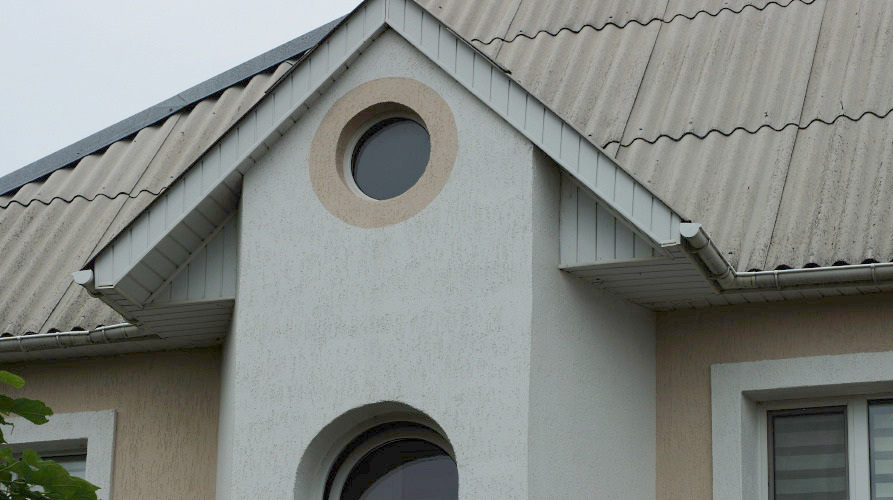 Nahaufnahme eines Hausdachs mit hellgrauen Wellblechpaneelen und einem kleinen Rundfenster im Giebelbereich der oberen Wand. 